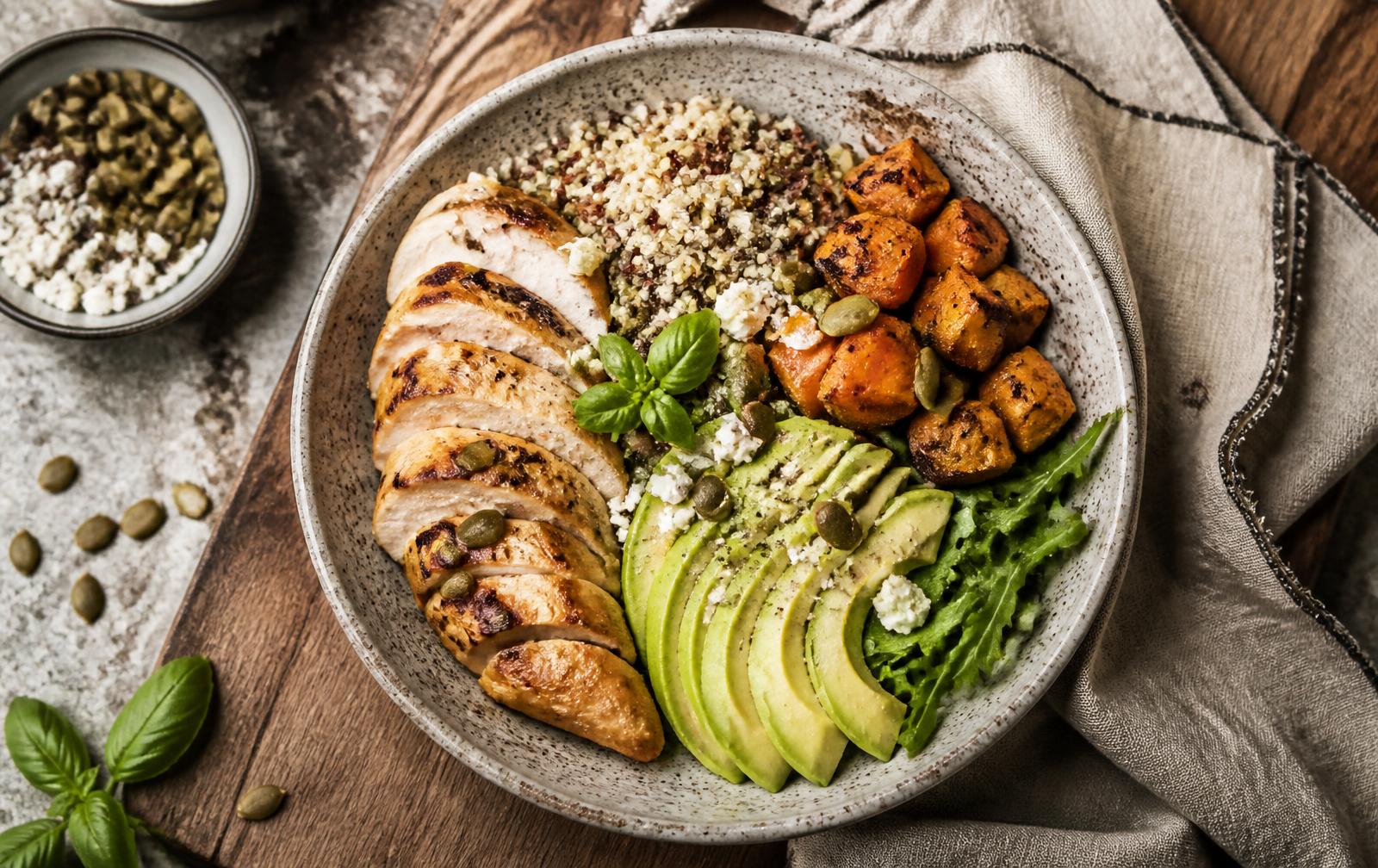 Kom met gegrilde kip, quinoa, avocado en geroosterde groenten op een houten tafel
